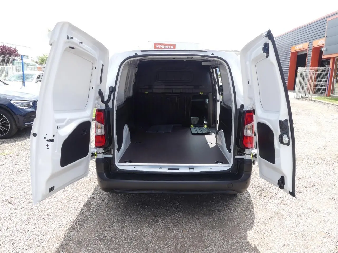 Vue arrière d'un Peugeot Partner blanc 2025 avec portes arrière ouvertes et plancher bois antidérapant visible dans la zone de chargement.