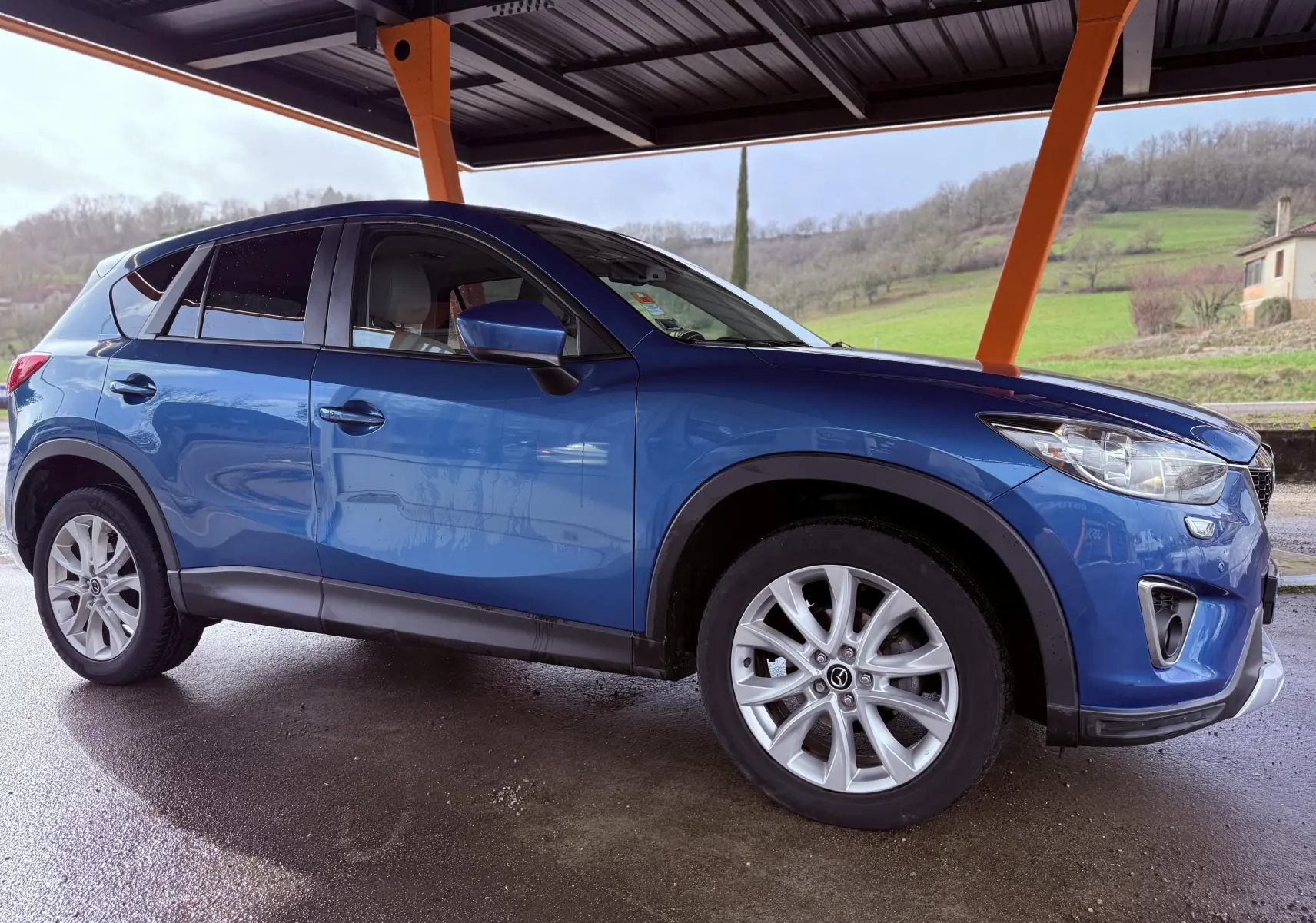 Vue de profil côté gauche d'un Mazda CX-5 bleu 2013 sous abri, avec jantes alliage et détails noirs contrastants.