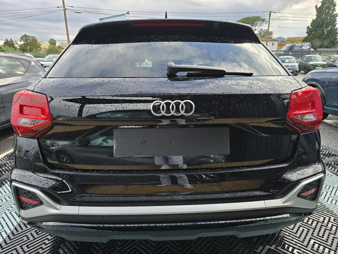 Vue arrière d'un Audi Q2 noir mythic avec feux rouges et lunette arrière surteintée sous la pluie.