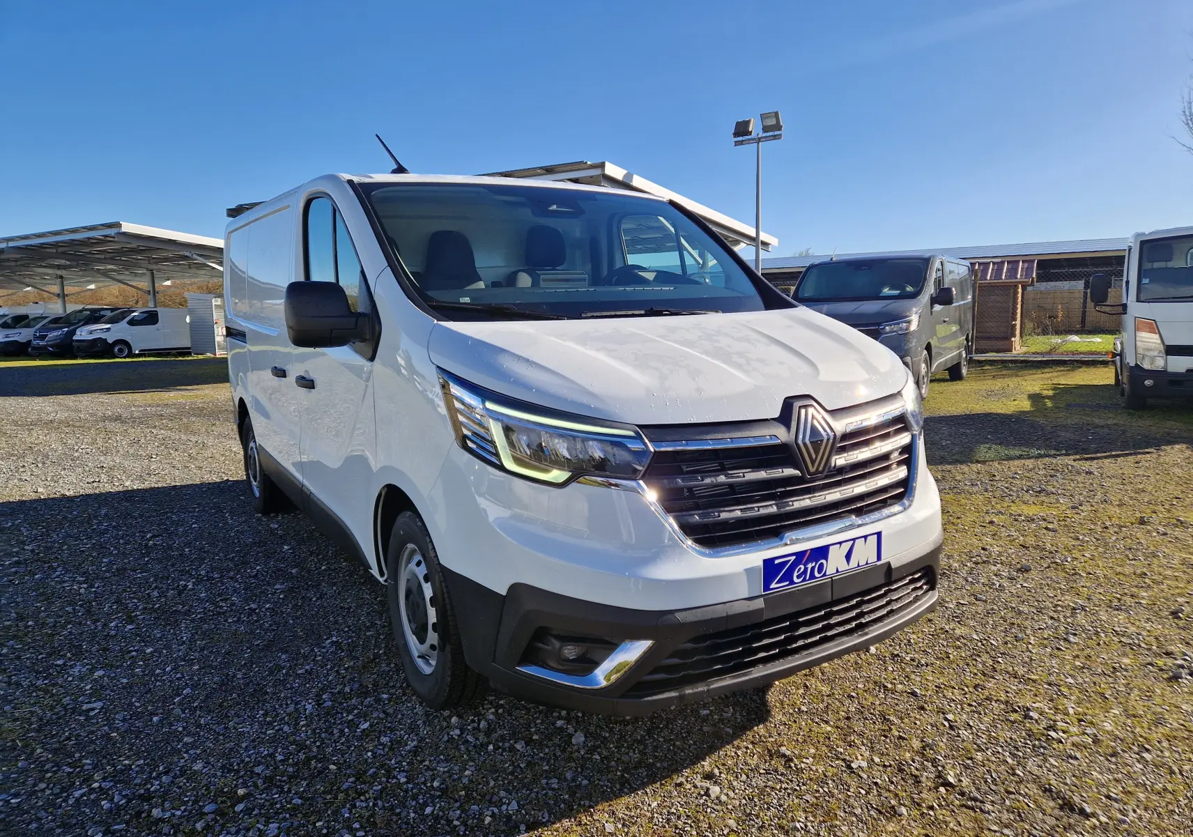 Renault Trafic Fourgon blanc vu en 3/4 avant droit, avec calandre noire et phares LED allumés, sur parking extérieur.