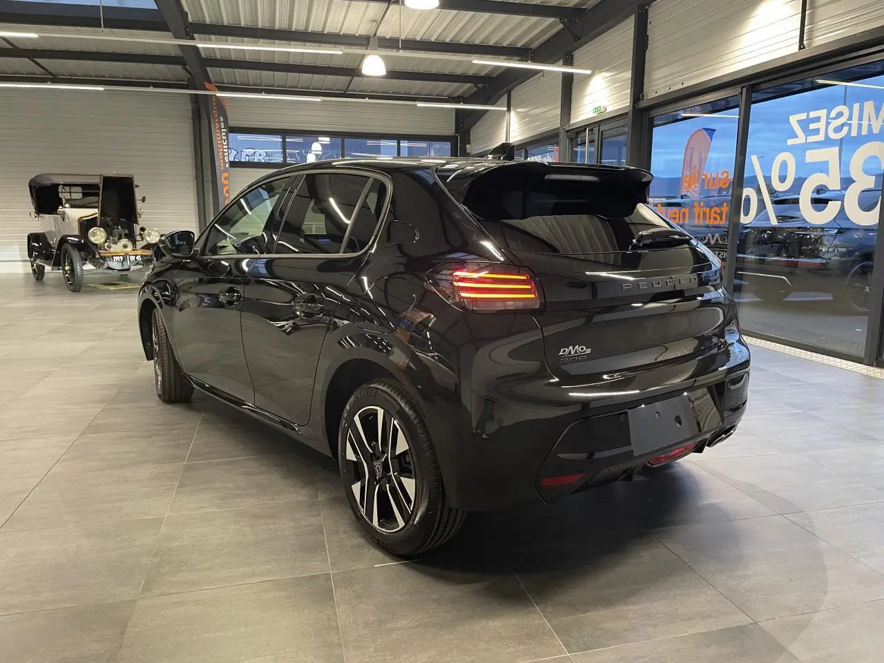 Peugeot 208 noire métallisée vue 3/4 arrière droit en intérieur avec feux arrière allumés et jantes alliage.