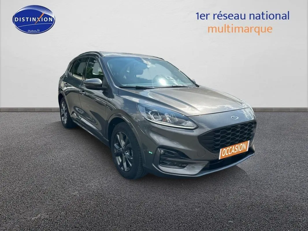 Ford Kuga gris foncé en 3/4 avant droit, avec jantes alliage 18 pouces et barres de toit visibles.