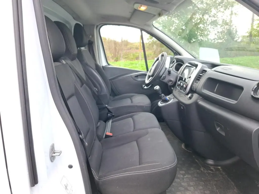 Vue intérieure côté conducteur du Renault Trafic Fourgon blanc, montrant les sièges noirs et le tableau de bord moderne.