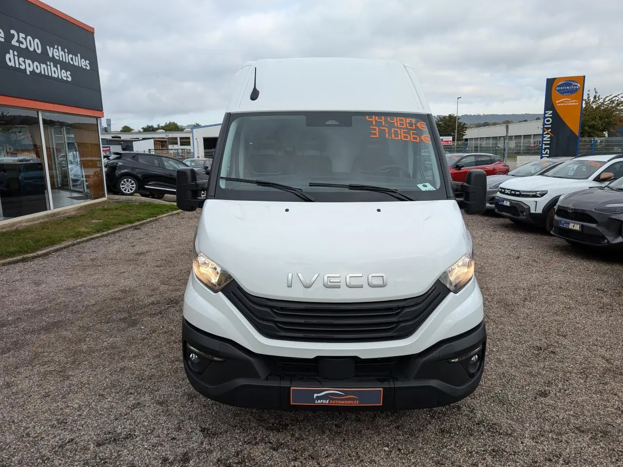 Vue frontale d'un utilitaire Iveco Daily blanc avec calandre noire et phares allumés sur un parking.