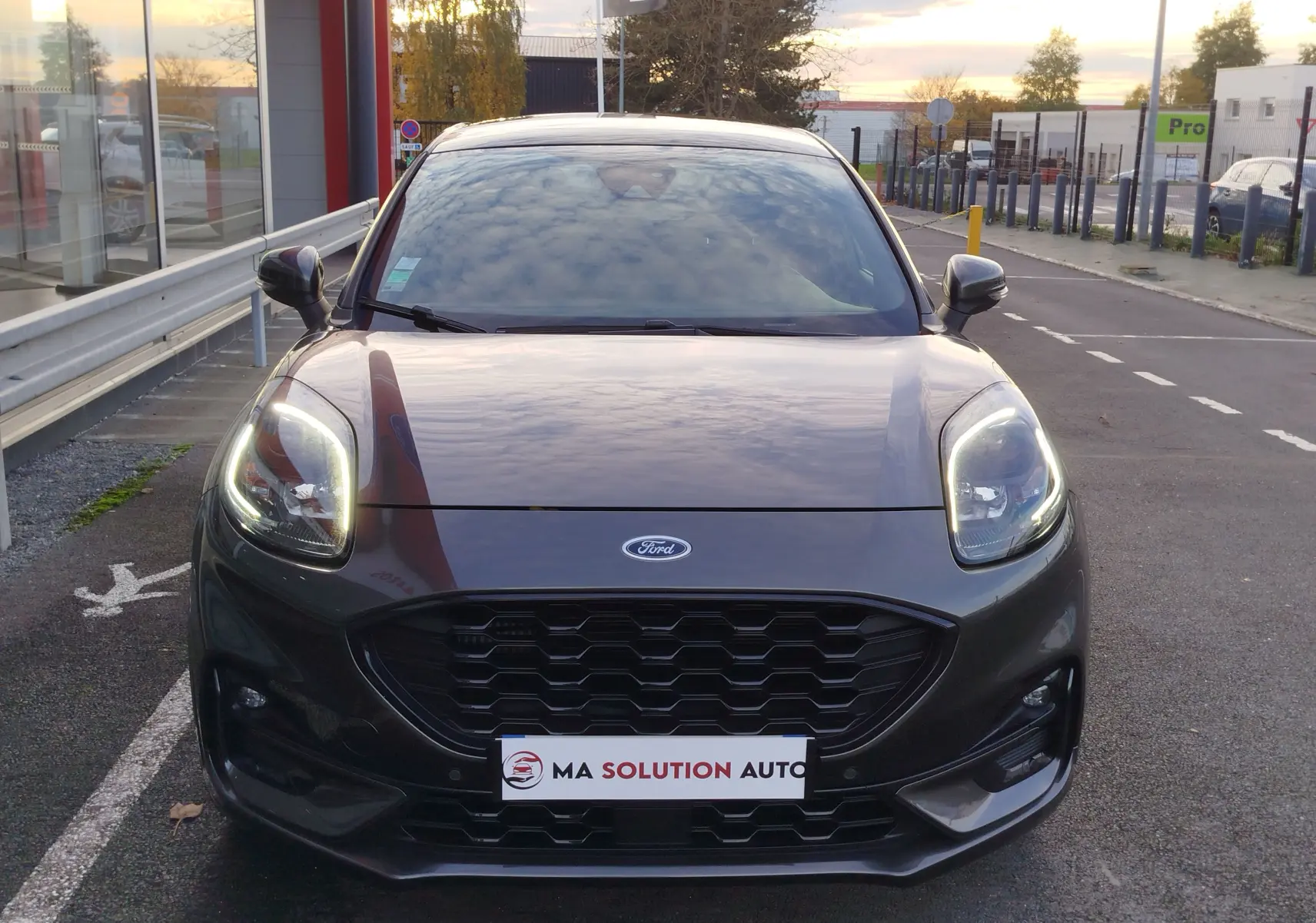 Vue frontale d'une Ford Puma 1.0 EcoBoost grise métallisée avec phares allumés et calandre noire hexagonale.