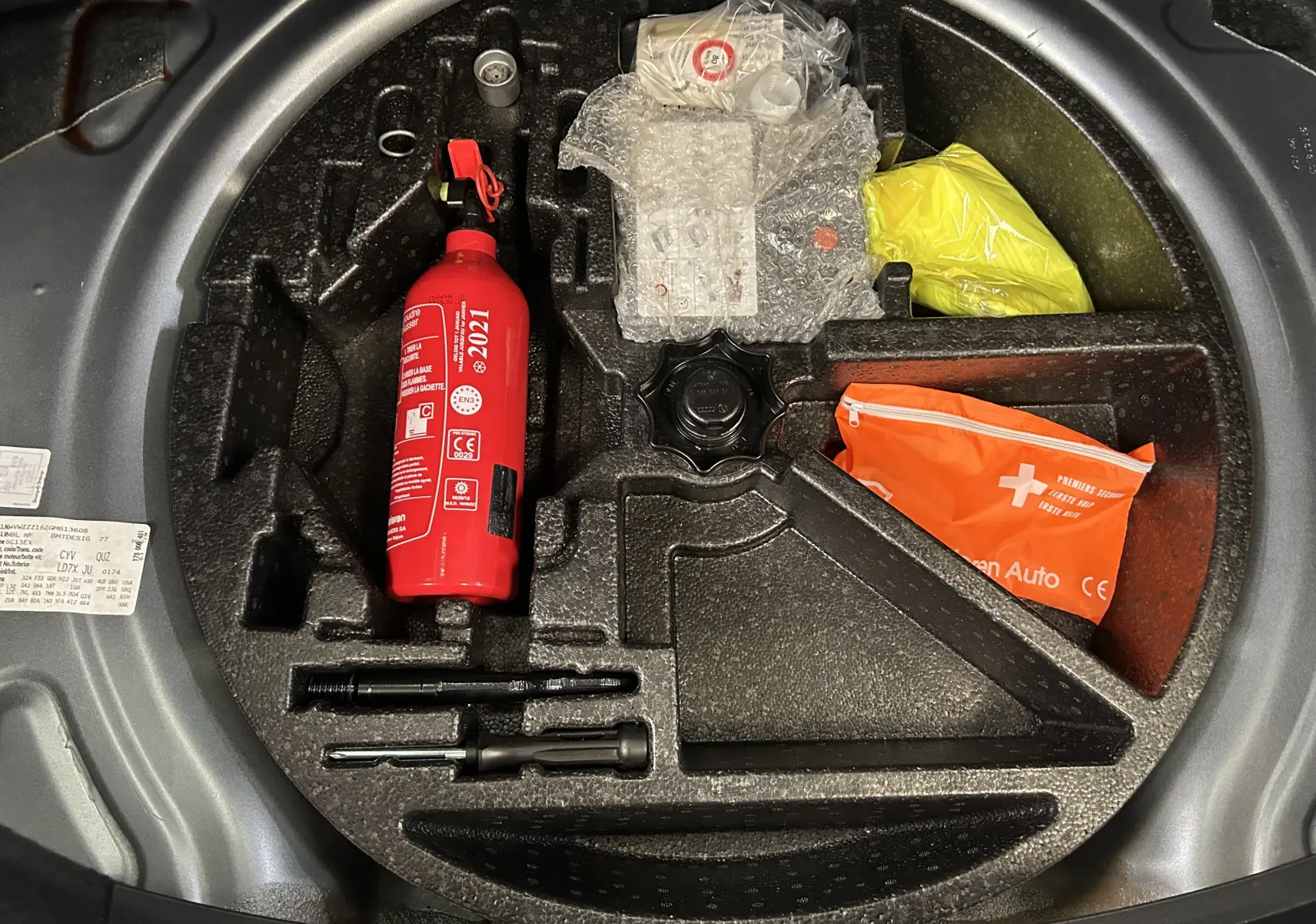 Vue plongeante sur le coffre de la Volkswagen Coccinelle gris, montrant le rangement avec extincteur rouge et trousse de secours orange.