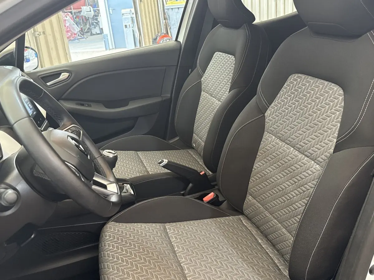Vue intérieure côté conducteur de la Renault Clio blanche 2023, montrant les sièges avant en tissu bicolore et le volant multifonction.
