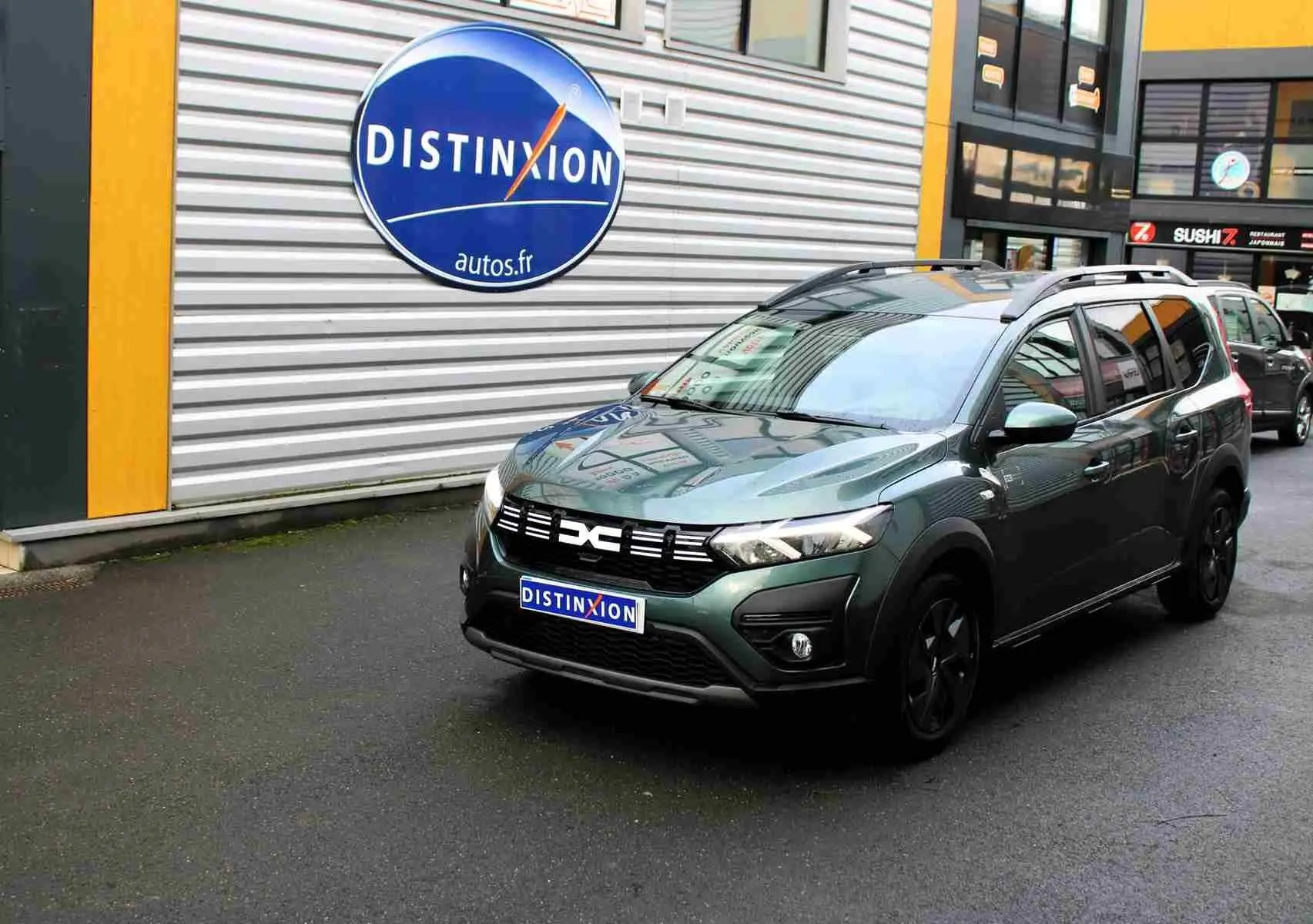 Dacia Jogger vert cèdre métal en 3/4 avant droit, avec calandre noire et barres de toit visibles.