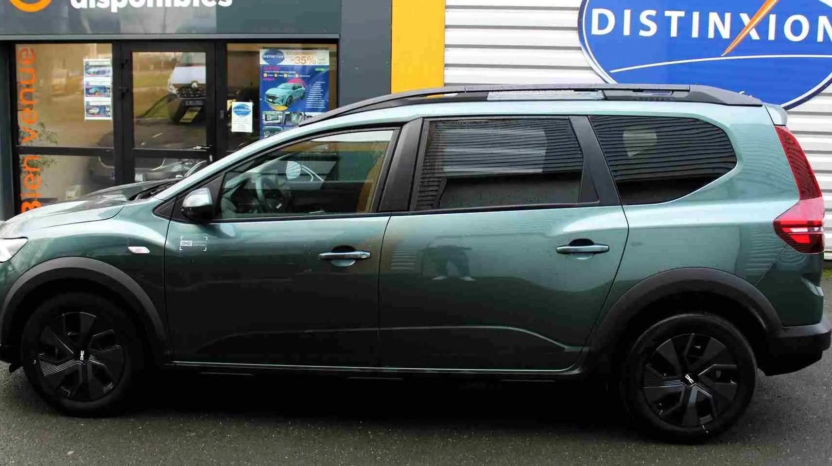 Vue de profil côté gauche d'une Dacia Jogger 2025 vert cèdre métal avec jantes noires et barres de toit.