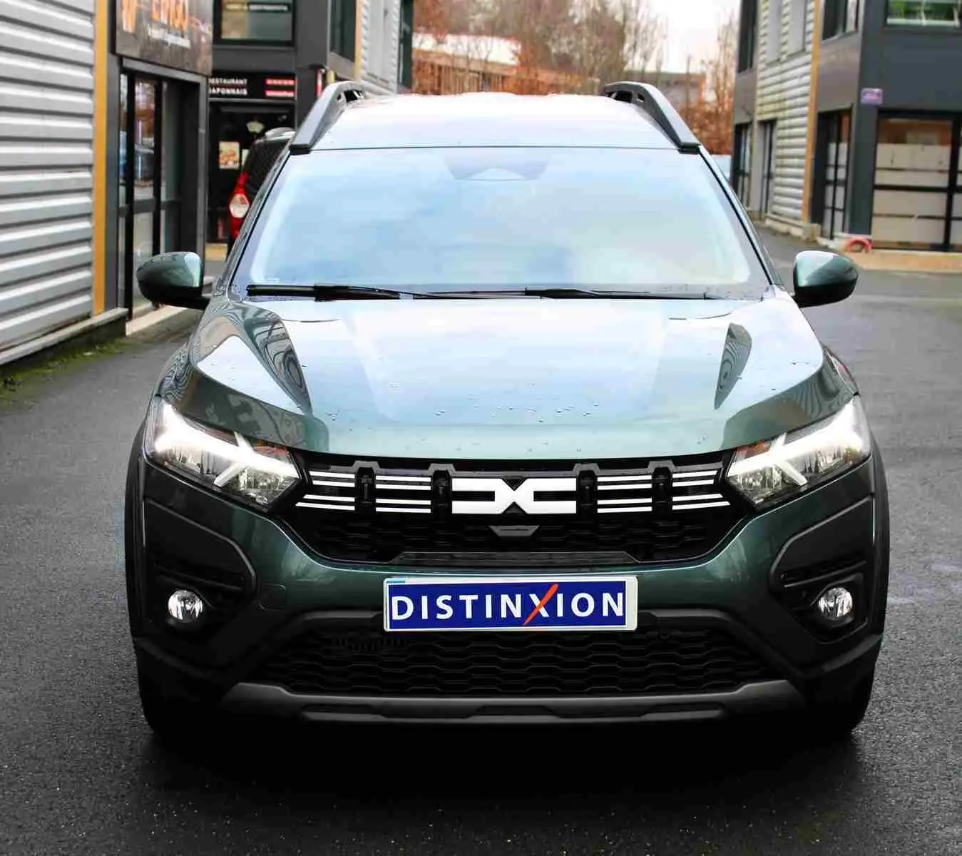 Vue avant d'un Dacia Jogger vert cèdre métal avec calandre noire et phares allumés sur fond urbain.