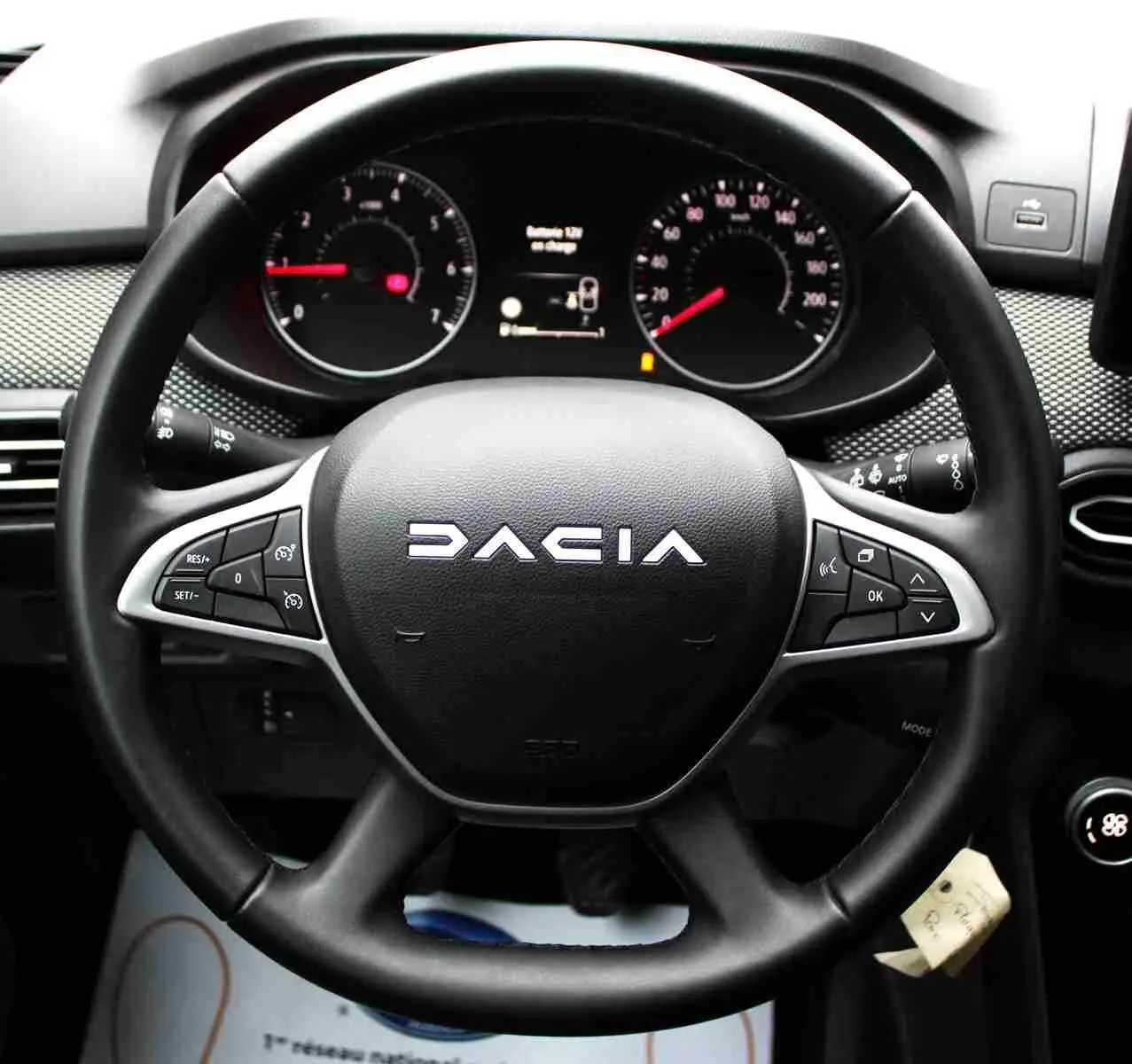 Vue rapprochée du volant noir DACIA avec commandes intégrées, tableau de bord et affichage numérique en arrière-plan.