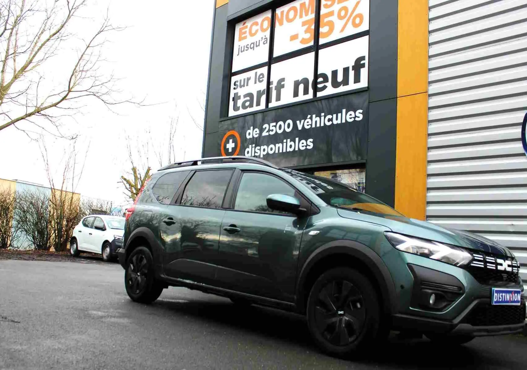 Dacia Jogger vert cèdre métal en vue 3/4 avant droit, stationné devant un bâtiment avec panneau promotionnel.