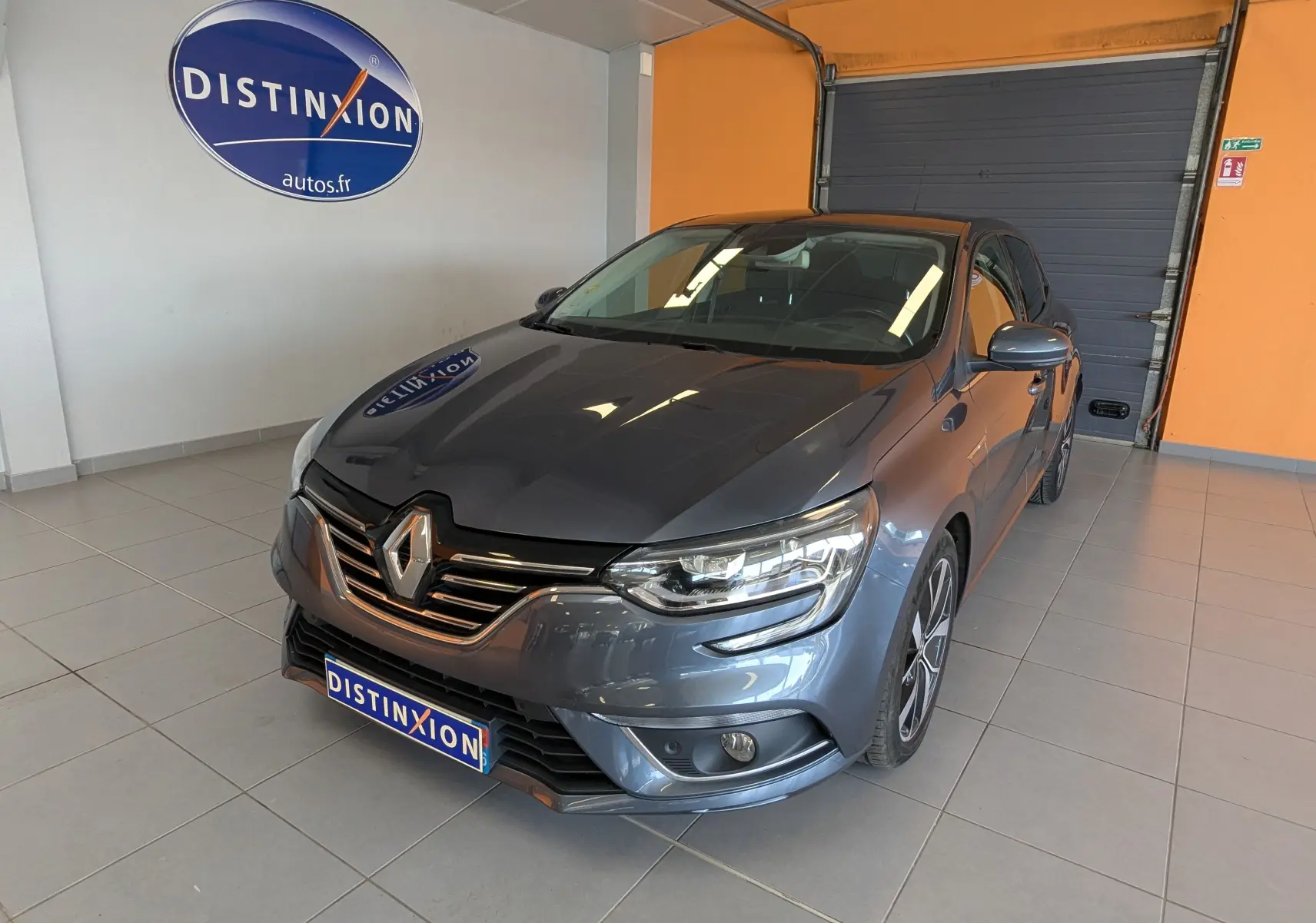 Renault Mégane IV gris en 3/4 avant droit, avec calandre chromée et jantes alliage bicolores en showroom.