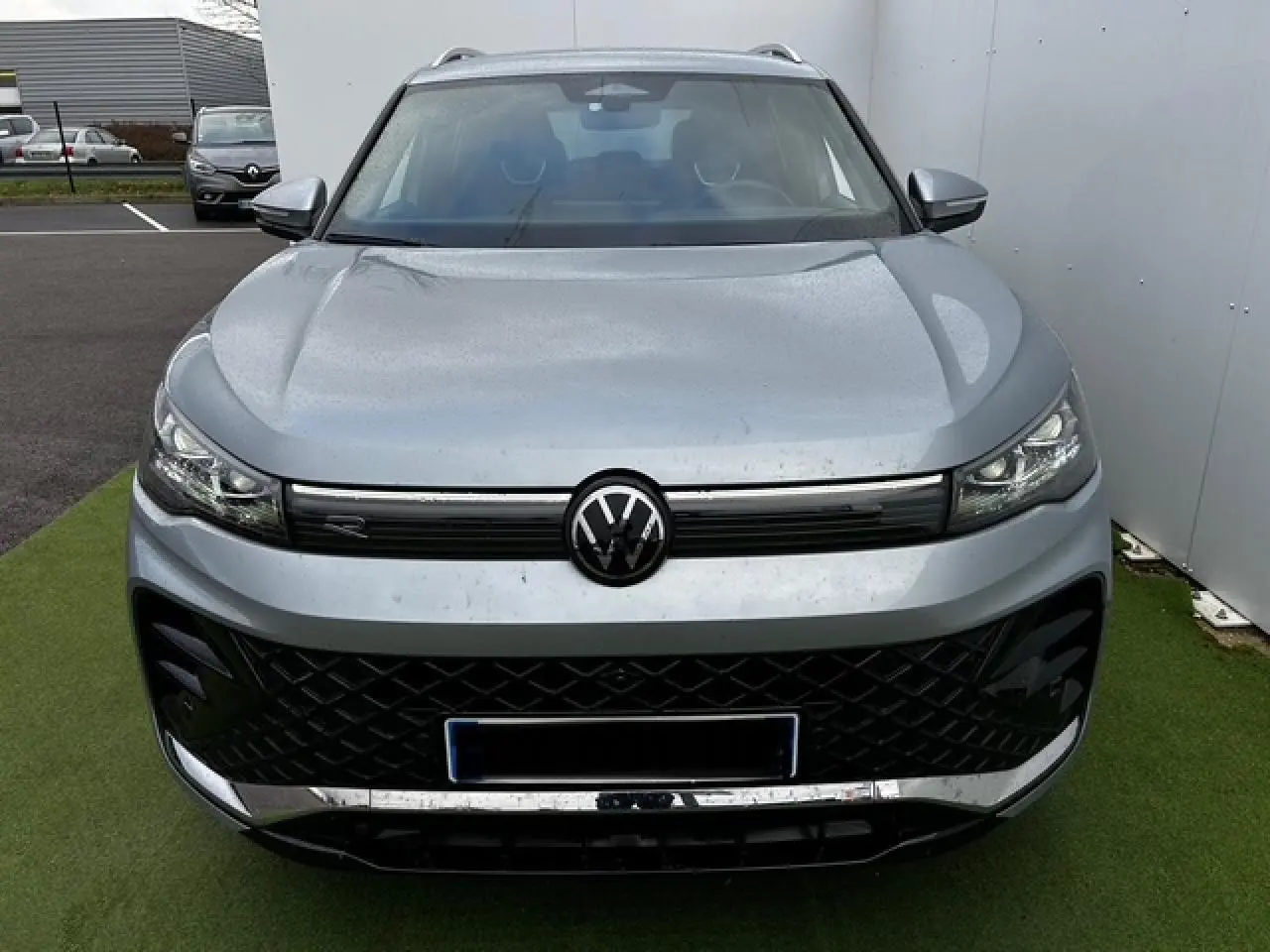 Vue frontale d'un Volkswagen Tiguan III gris clair avec calandre noire et logo VW central sur fond urbain.