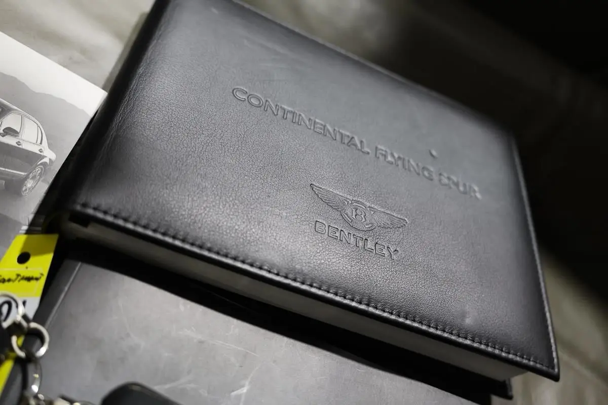 Image montrant un carnet d'entretien en cuir noir embossé Bentley Continental Flying Spur, posé sur une surface grise.