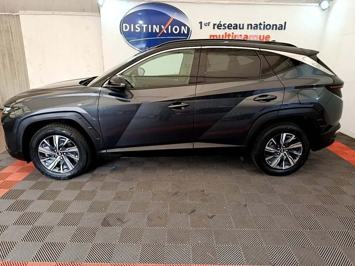 Vue de profil côté gauche d'un Hyundai Tucson 1.6 Hybrid 230 Creative gris foncé avec jantes alliage en intérieur showroom.