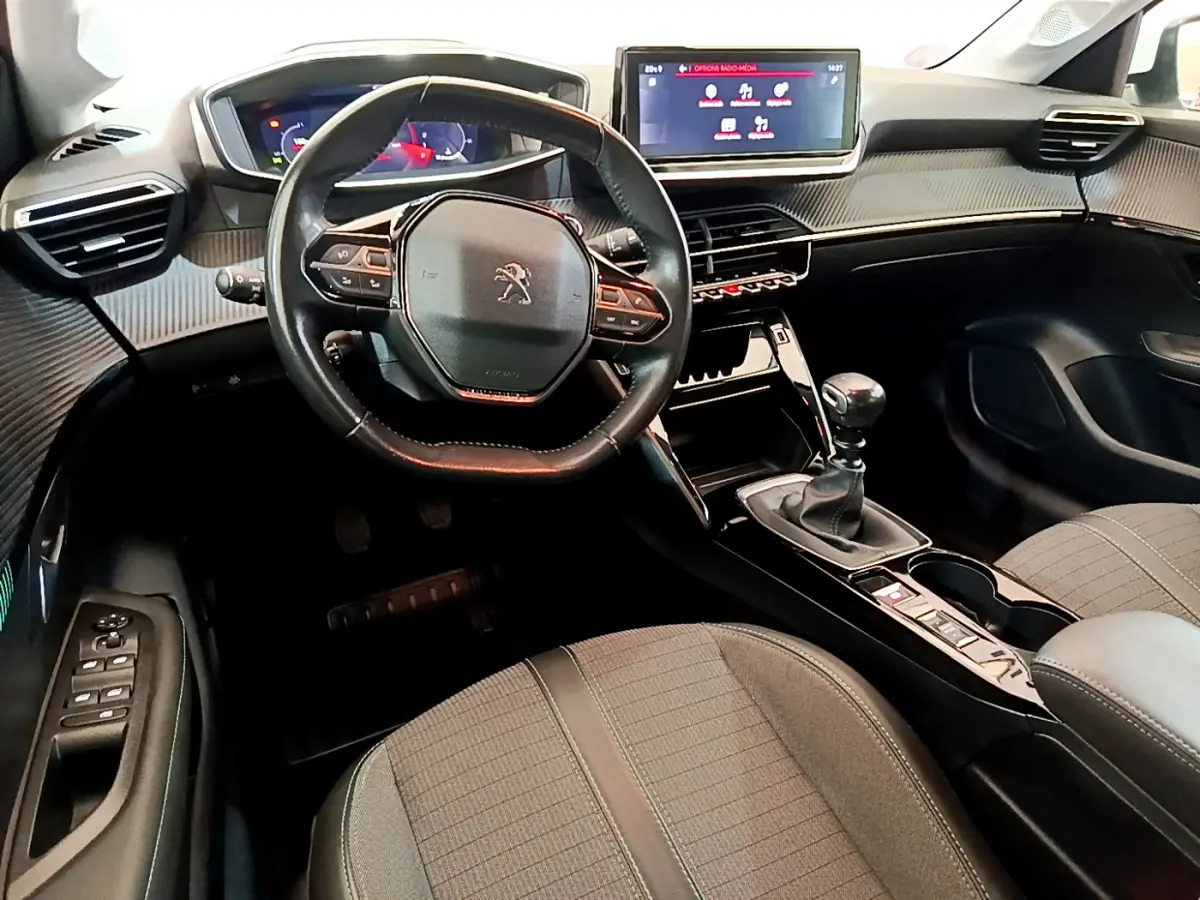 Vue intérieure avant du Peugeot 208 blanc, volant cuir compact, écran tactile 10 pouces et boîte manuelle visible.