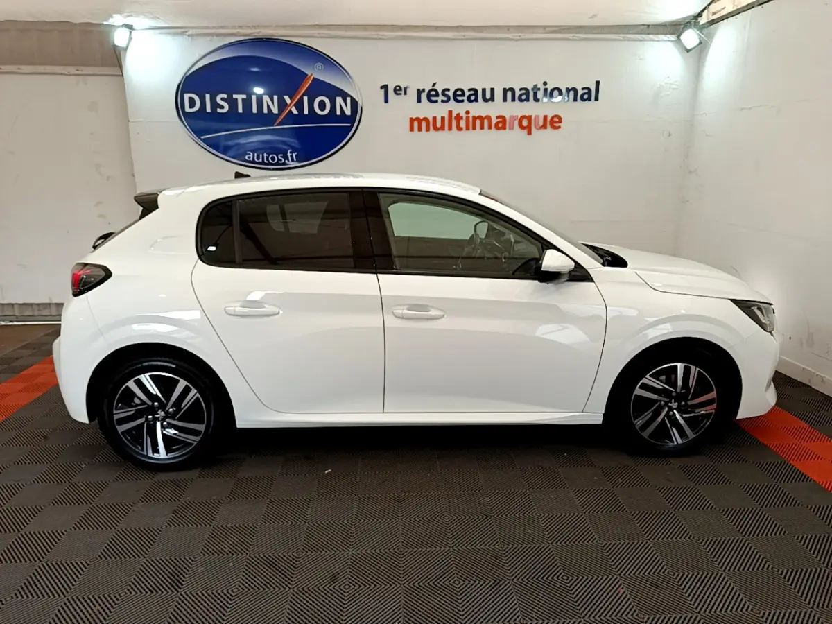 Profil côté gauche d'une Peugeot 208 blanche PureTech 100 S&S Allure Business, en intérieur sur sol damier noir et rouge.