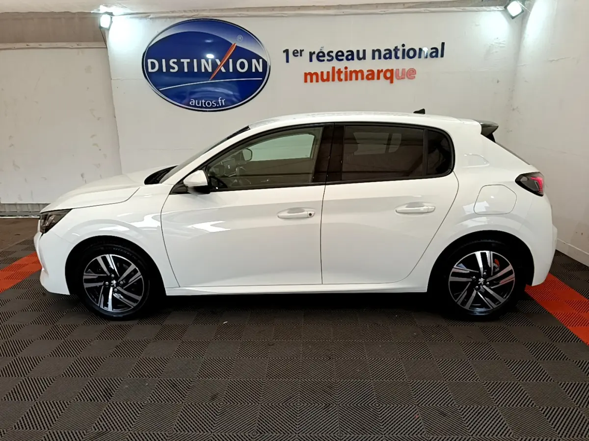 Profil côté gauche d'une Peugeot 208 blanche PureTech 100 S&S Allure Business, en intérieur sur sol damier noir et rouge.