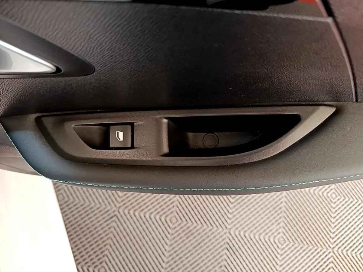 Gros plan sur la commande de lève-vitre côté gauche intérieur d'une Peugeot 208 blanche avec surpiqûres bleues.