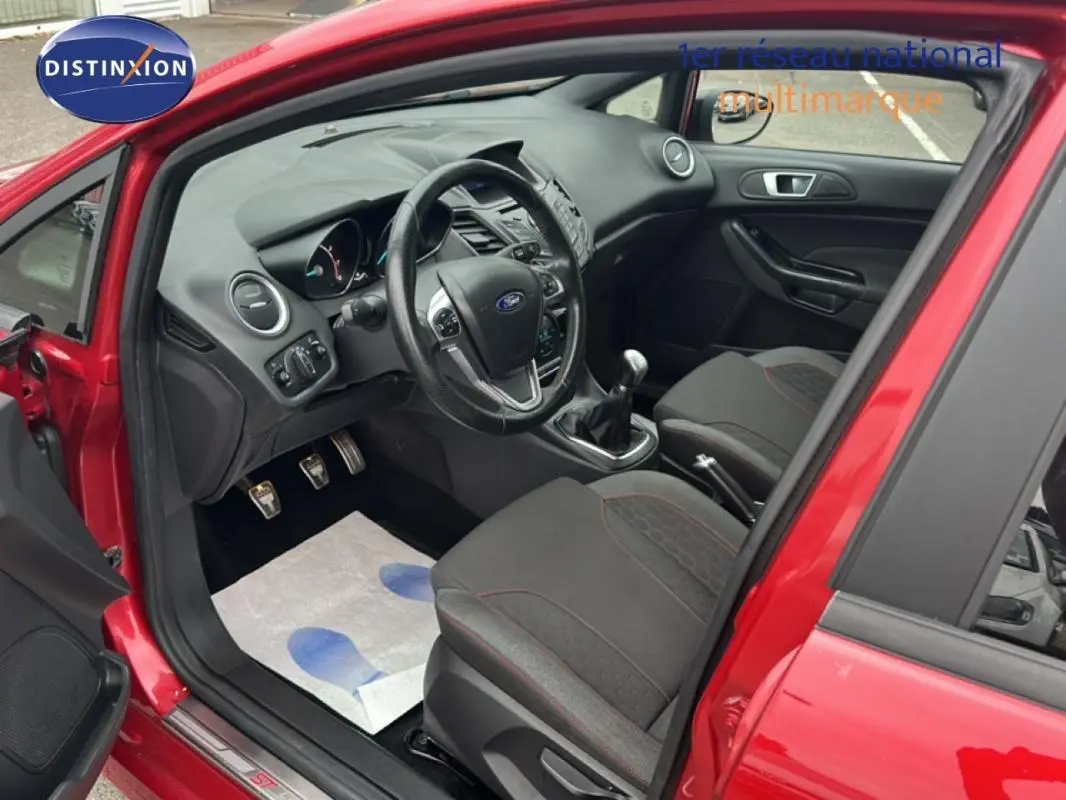 Vue intérieure côté conducteur d'une Ford Fiesta rouge 2017 avec volant cuir, boîte manuelle et sièges tissu noirs.