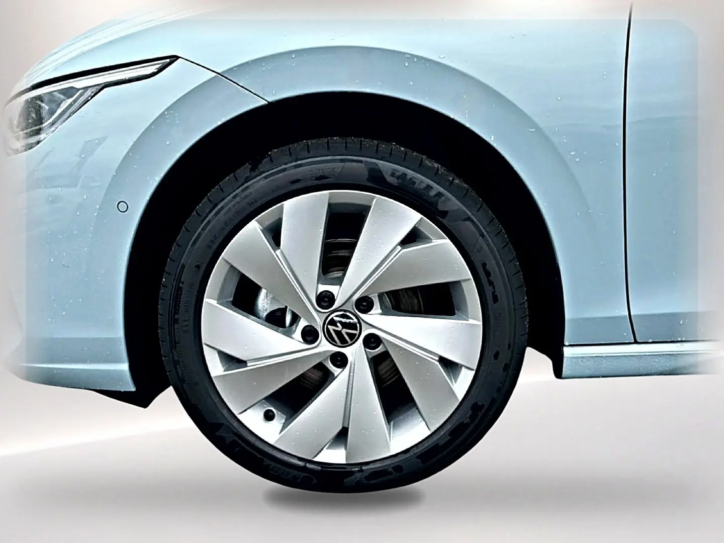 Gros plan sur la roue avant gauche d'une Volkswagen Golf bleu glacier avec jante alu 17 pouces et partie du phare avant visible.