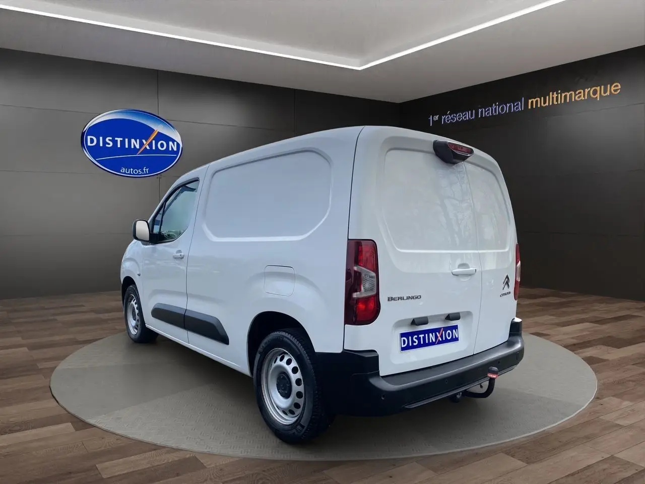 Vue 3/4 arrière droite d'un utilitaire Citroën Berlingo blanc avec hayon fermé et attelage visible en intérieur showroom.