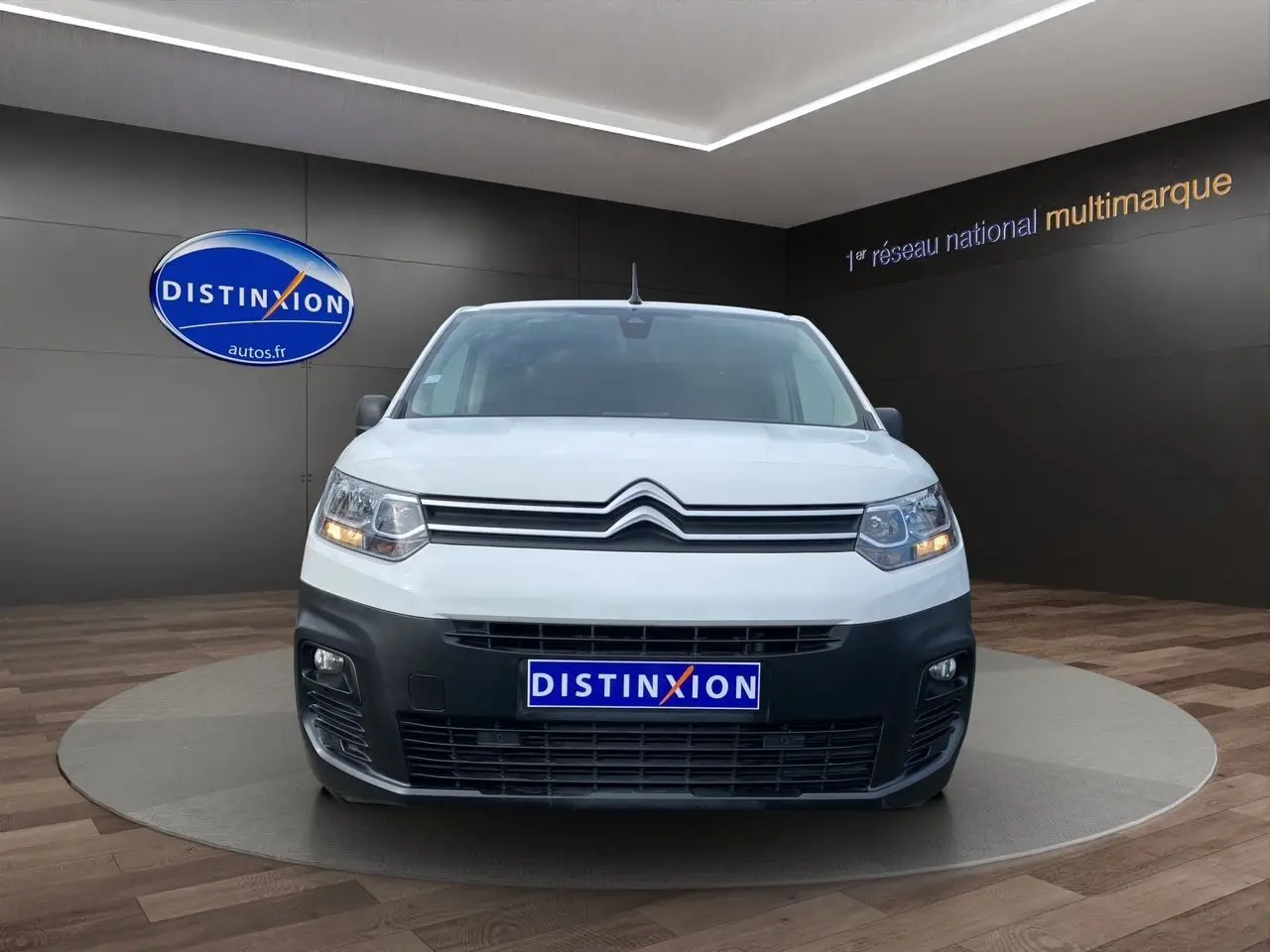 Vue de face d'un Citroën Berlingo blanc avec pare-chocs noir et feux allumés en showroom.