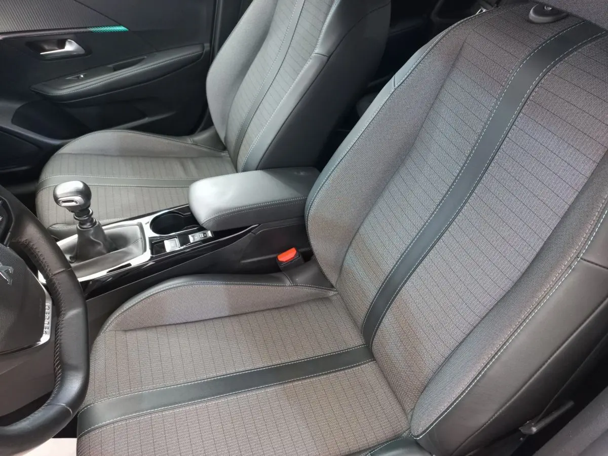 Vue intérieure côté conducteur de la Peugeot 208 PureTech 100 S&S Allure 2021, sièges tissu gris avec surpiqûres noires.