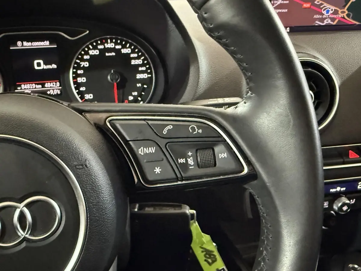 Gros plan sur le volant cuir noir de l'Audi A3 2018 avec commandes audio et tableau de bord affichant la navigation.