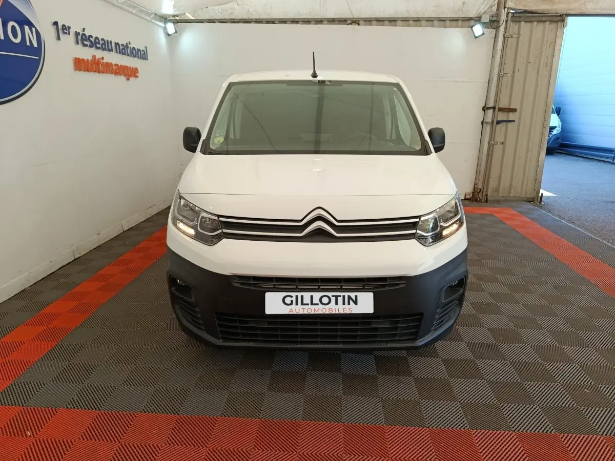 Vue frontale d’un Citroën Berlingo blanc 2022 avec pare-chocs noir et phares allumés en intérieur showroom.