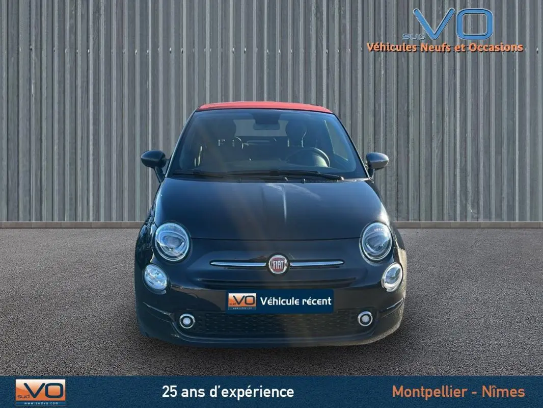 Vue frontale d'une FIAT 500C noire avec toit rouge, modèle 2024, affichée sur un parking devant un mur métallique.