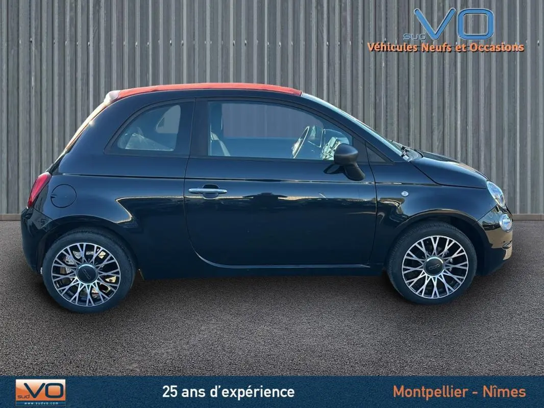 Profil côté gauche d'une FIAT 500C noire 2024 avec toit rouge et jantes stylisées, stationnée devant un mur métallique.