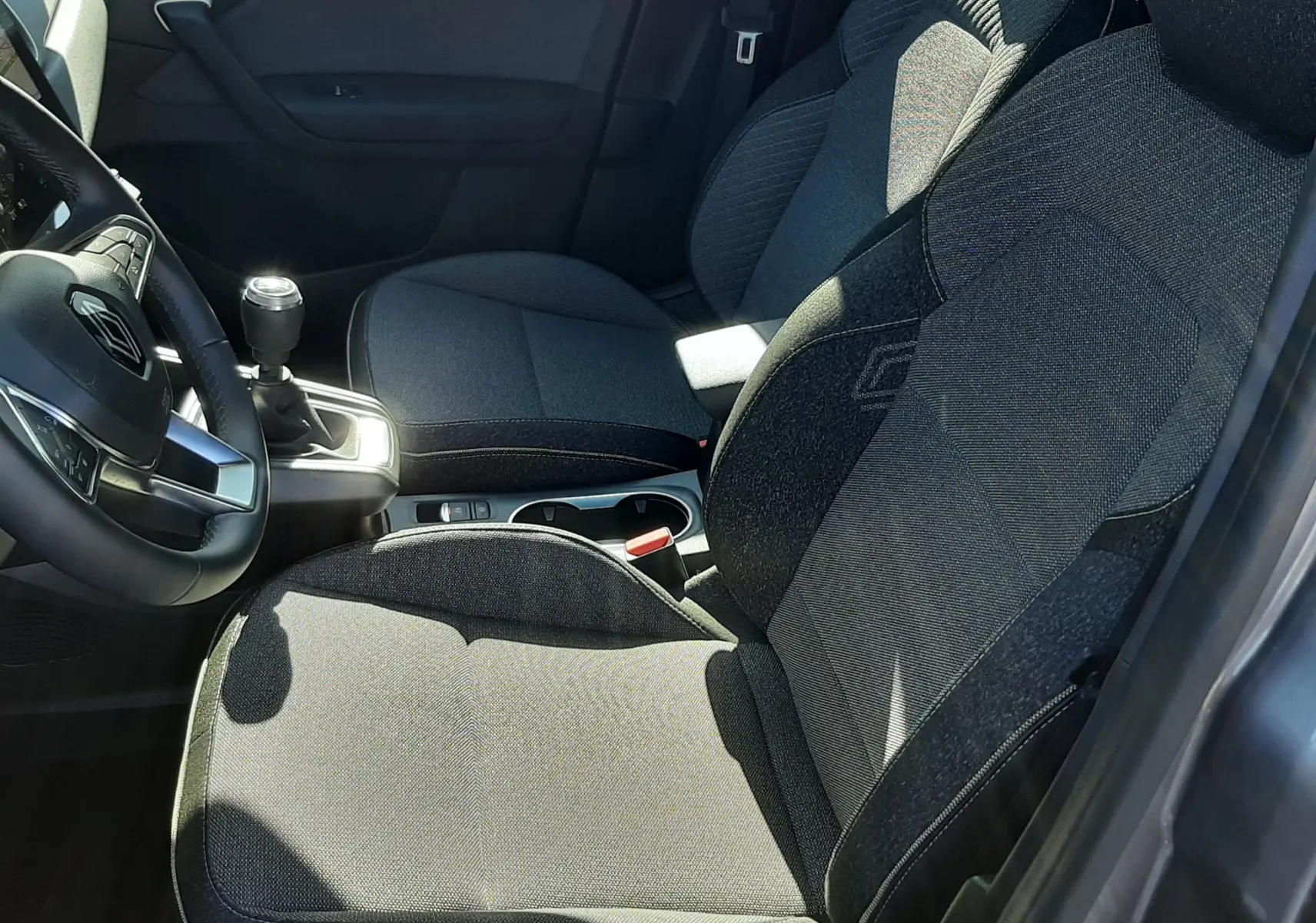 Vue intérieure côté conducteur du Renault Captur gris cassiopée 2025, sièges tissu gris avec logo brodé et levier de vitesse manuel.