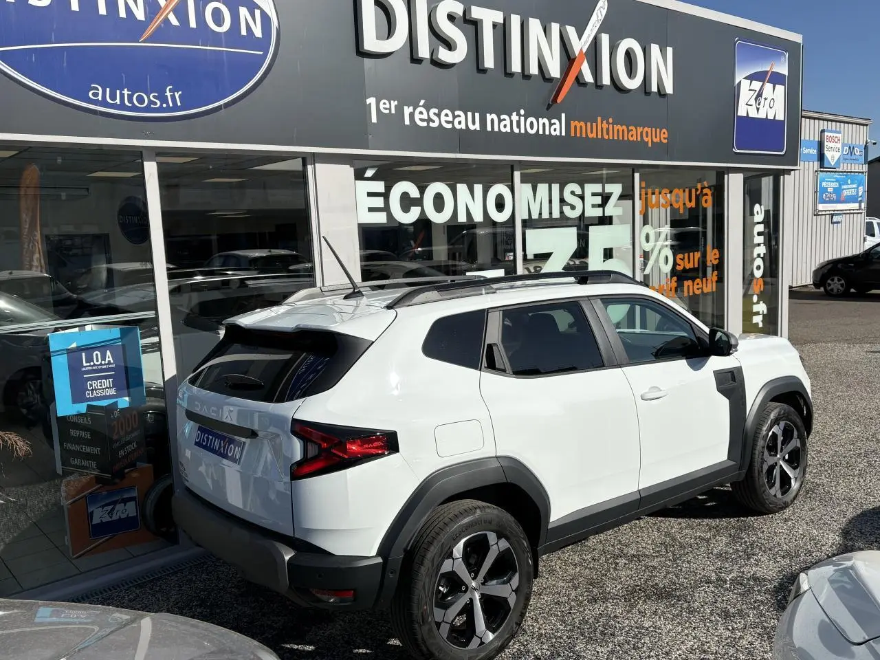 Vue 3/4 arrière droite d'un Dacia Duster blanc métallisé avec barres de toit noires et jantes alliage 18 pouces.