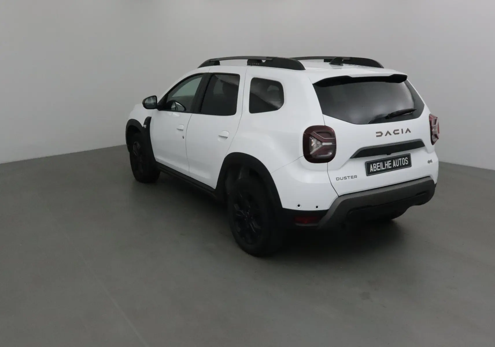 Vue 3/4 arrière droite du Dacia Duster 2024 blanc avec jantes noires et barres de toit noires dans un studio.