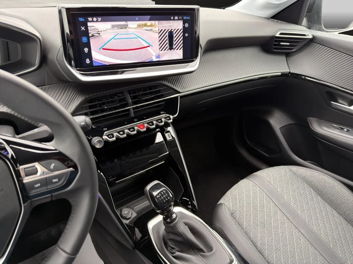 Vue intérieure côté conducteur de la Peugeot 208 gris clair, avec écran de caméra de recul et levier de vitesse manuel visible.