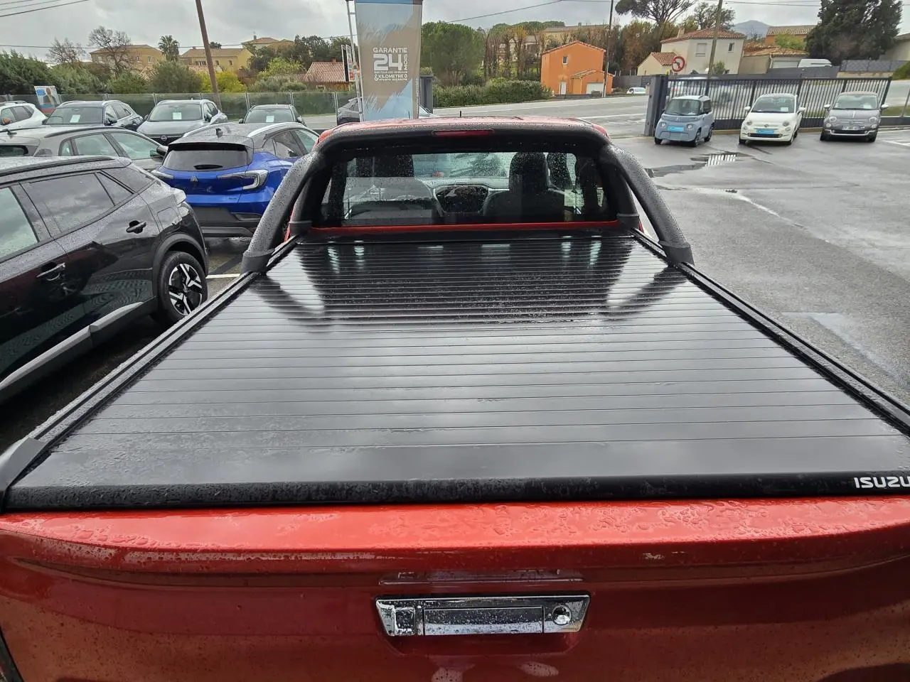 Vue arrière d'un pick-up Isuzu D-MAX orange métallisé avec benne couverte par un rideau rigide noir.
