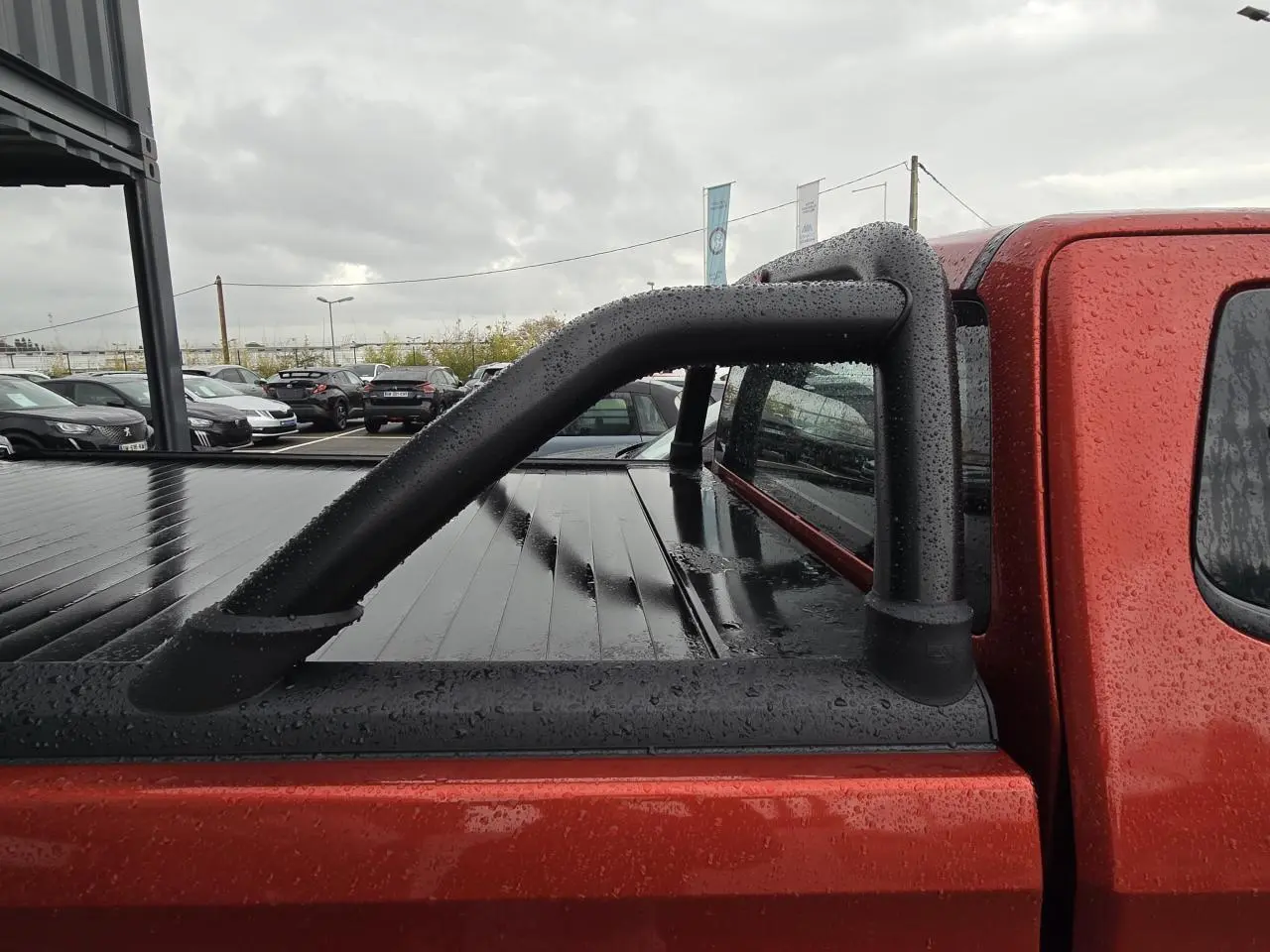 Vue latérale droite du pick-up Isuzu D-MAX orange métallisé avec sport bar noir et bac de benne fermé sous la pluie.
