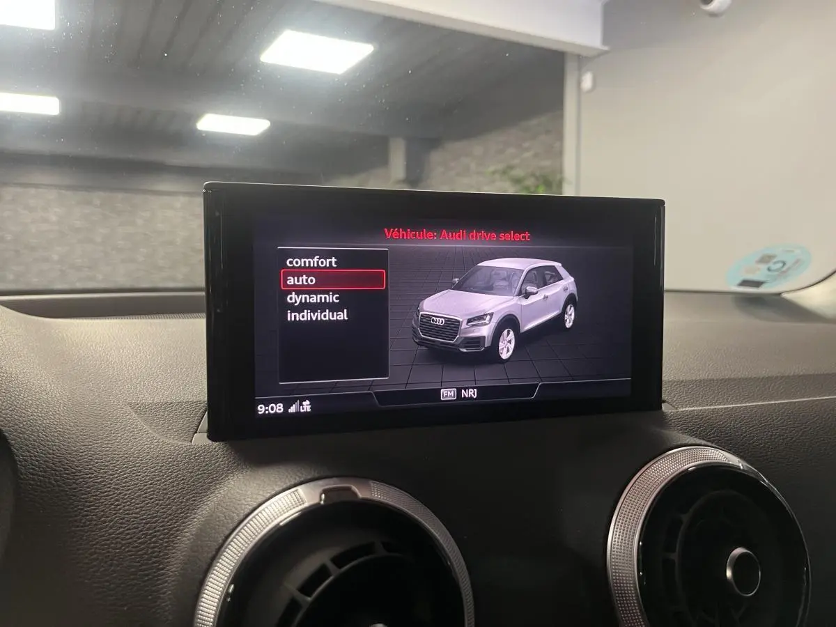 Écran central montrant un Audi Q2 gris en vue 3/4 avant avec options de mode de conduite sur fond noir.