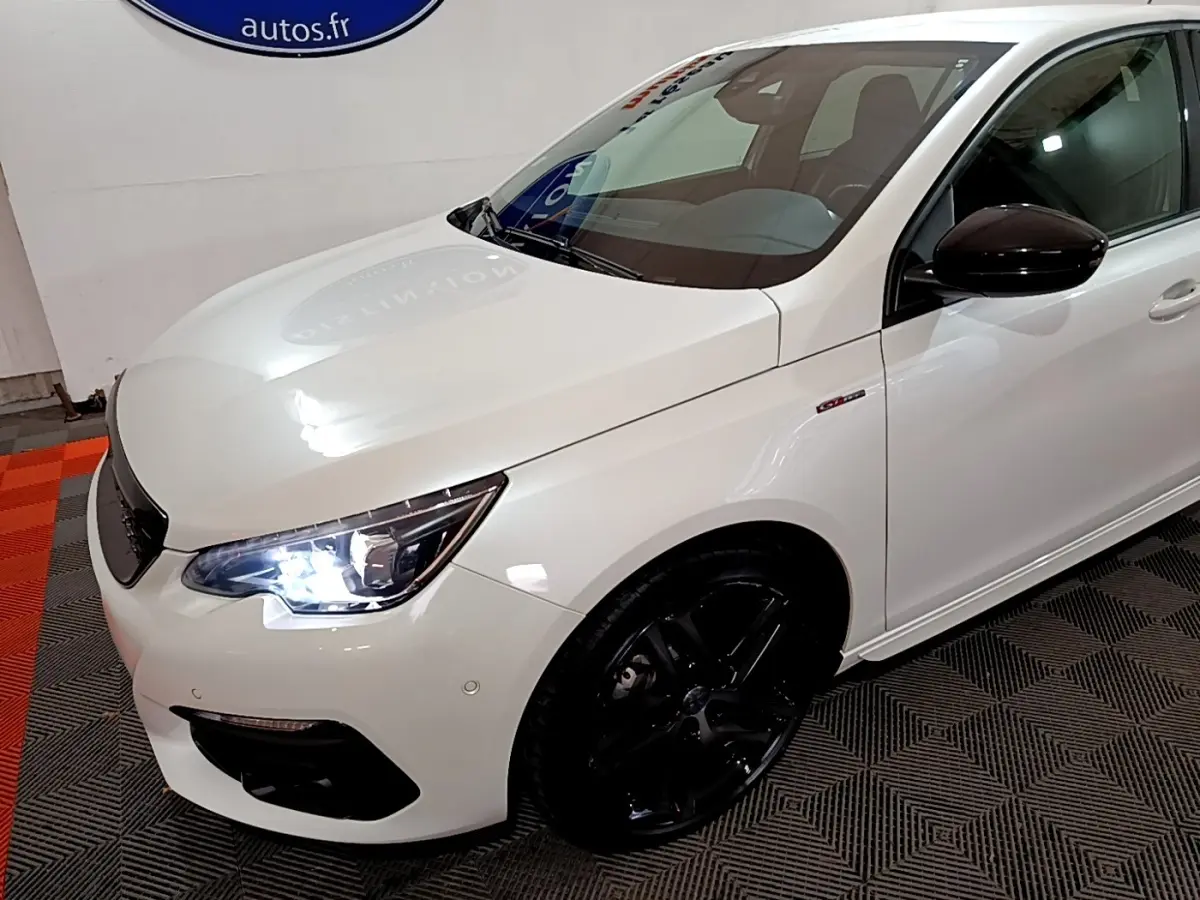 Vue 3/4 avant droit d'une Peugeot 308 blanche avec phares allumés et jantes noires brillantes.