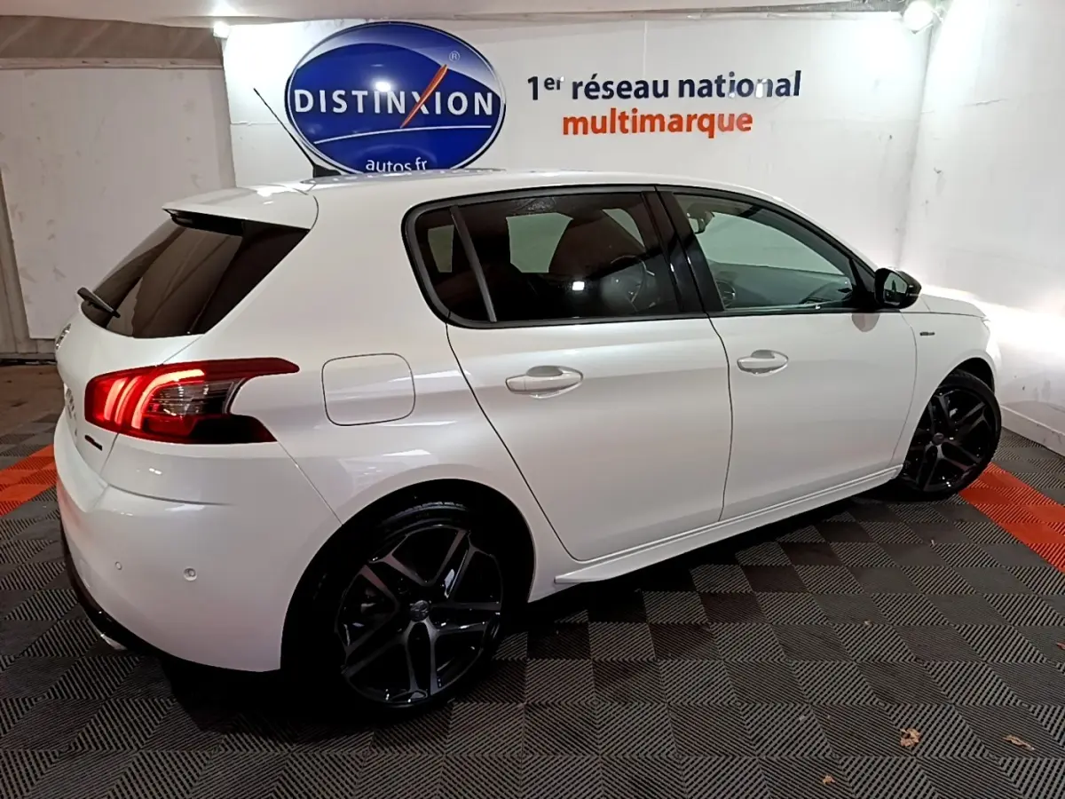 PEUGEOT 308 blanc vue côté droit arrière en intérieur, avec jantes noires et feux arrière allumés.
