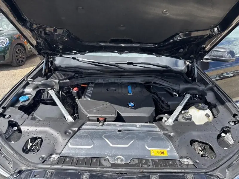 Vue avant du compartiment moteur ouvert d'un BMW X3 xDrive30e 2023 en Saphirschwarz métallisé, montrant le moteur hybride.