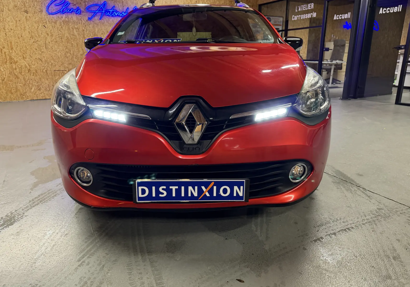 Vue frontale d'une Renault Clio Estate IV rouge avec feux diurnes allumés et plaque Distinxion visible.