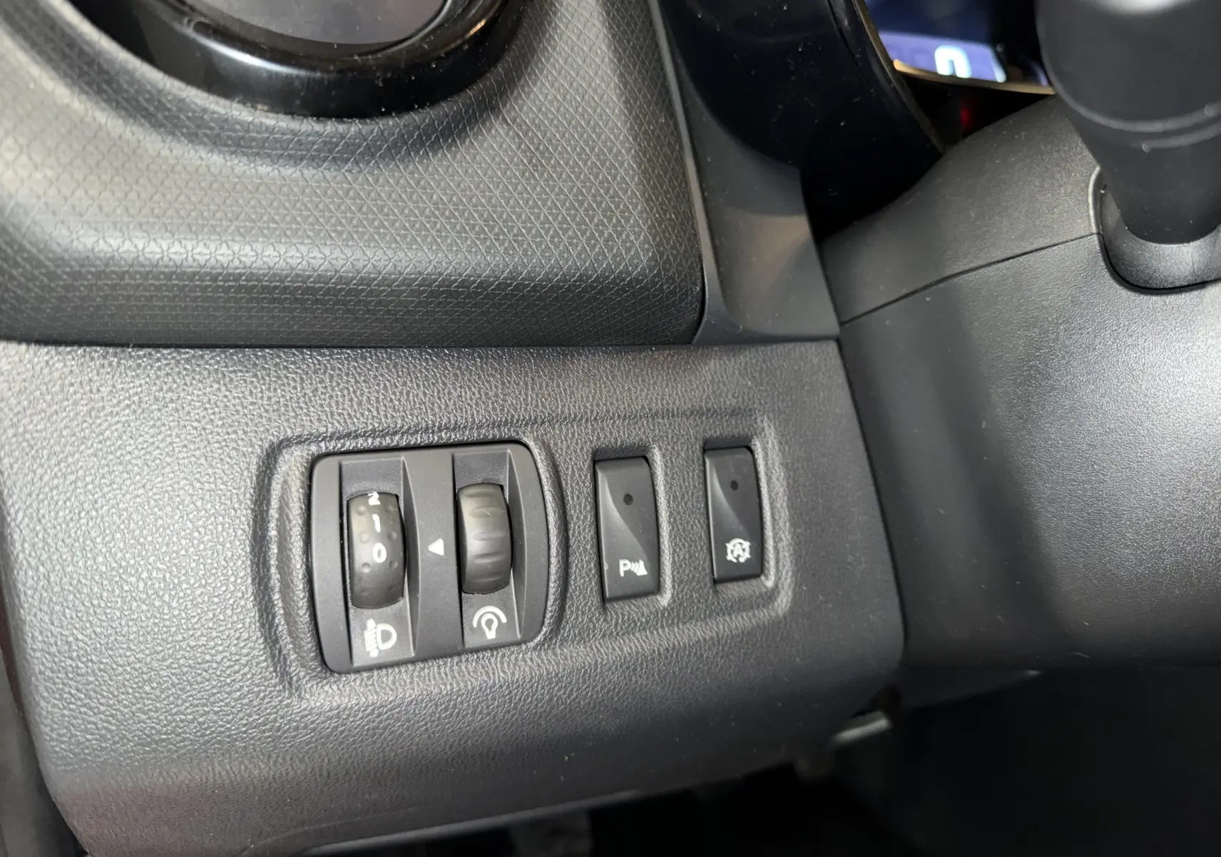 Gros plan sur les commandes de réglage des phares et aides à la conduite dans une Renault Clio Estate IV rouge, côté conducteur.