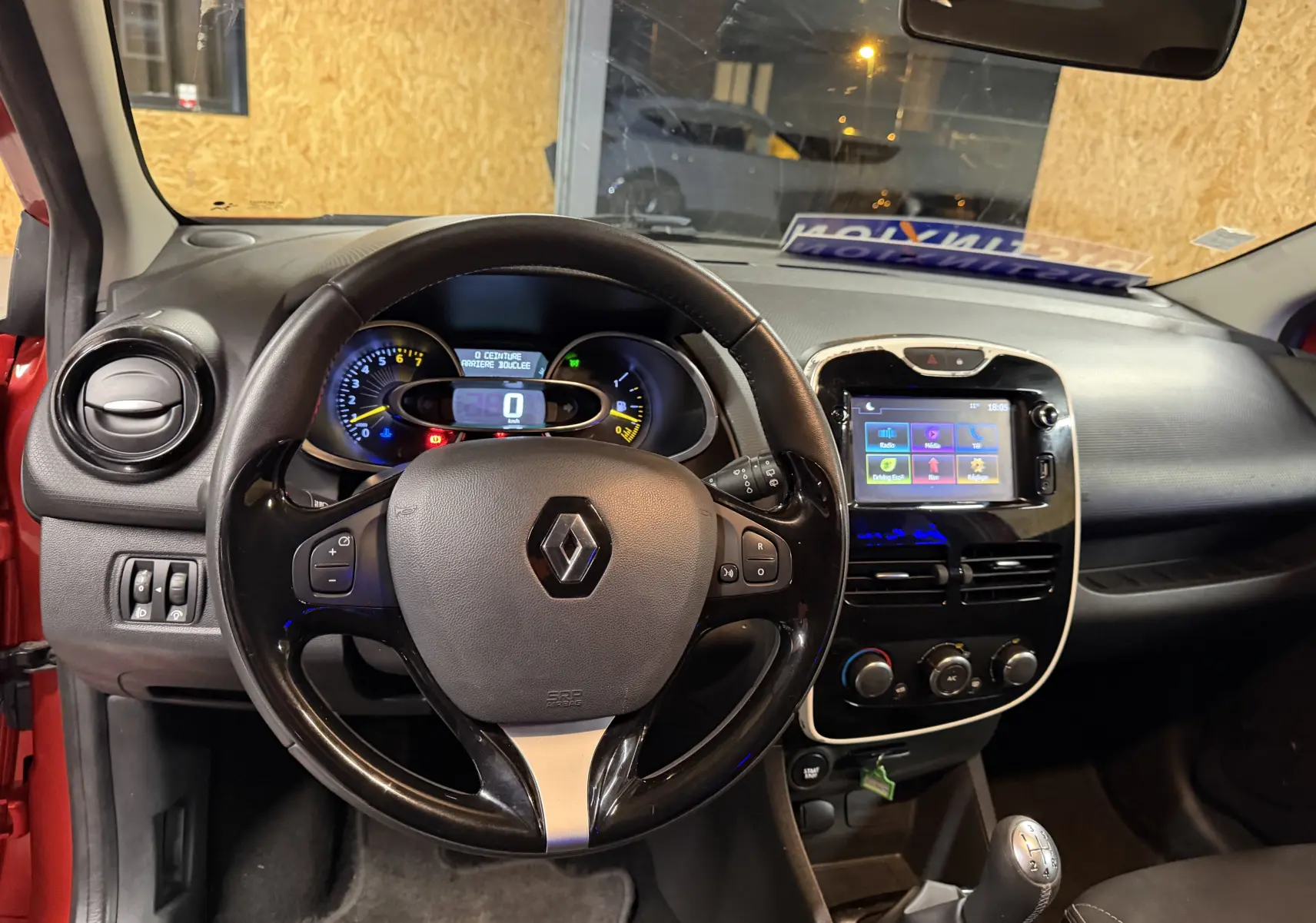 Vue intérieure du tableau de bord de la Renault Clio Estate IV rouge, avec volant, écran tactile et levier de vitesse manuel visibles.