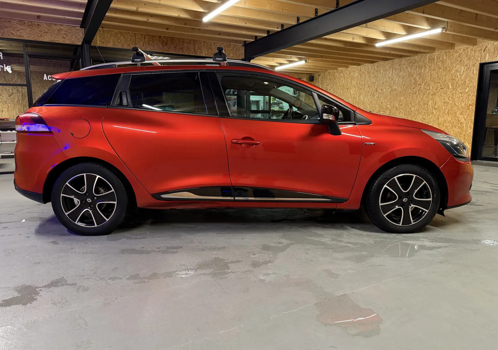 Profil côté gauche d'une Renault Clio Estate IV rouge 2015 avec barres de toit et jantes noires en intérieur garage.