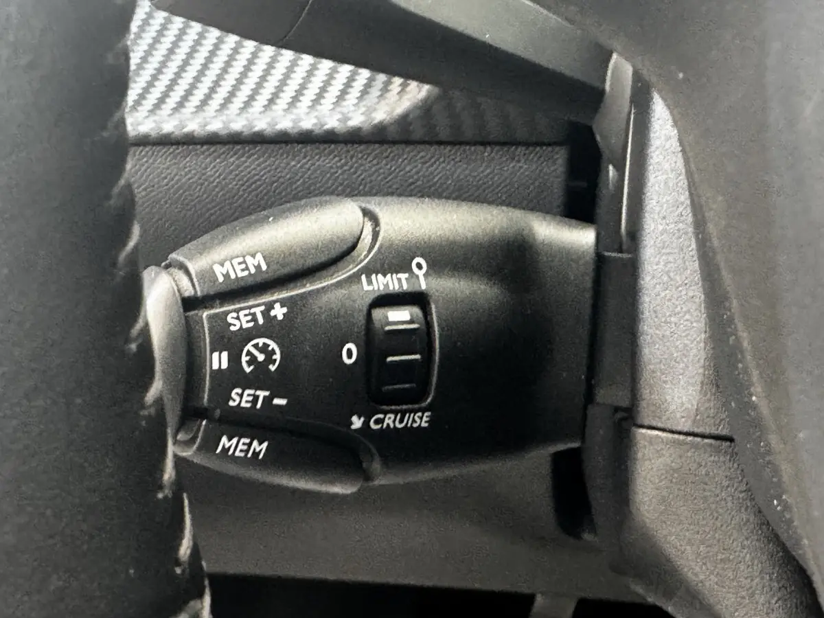 Gros plan sur la commande de régulateur de vitesse noire à gauche du volant d'une Peugeot 208 blanche.