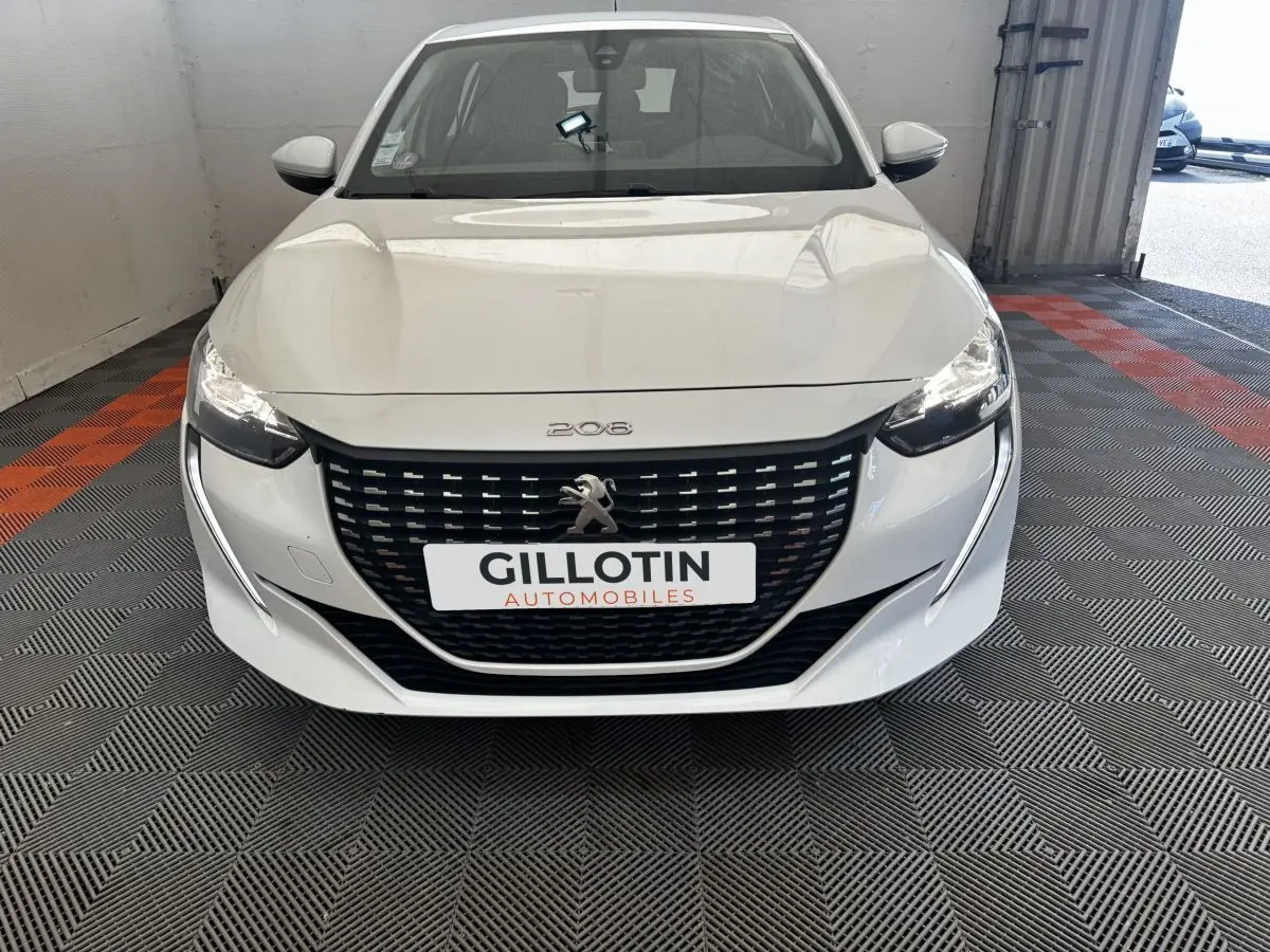 Vue avant d'une Peugeot 208 blanche avec calandre noire et phares allumés dans un garage.