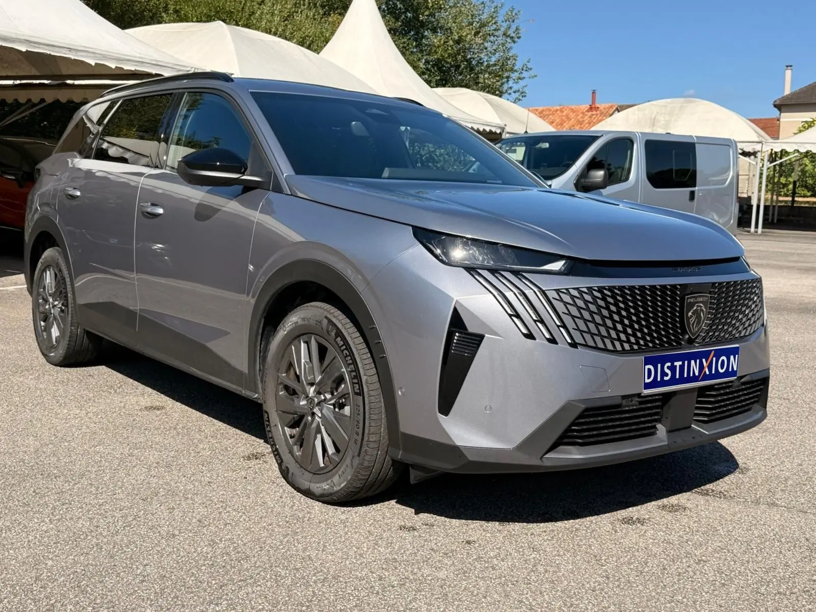 Peugeot 5008 gris Artense en 3/4 avant droit, avec calandre noire distinctive et jantes foncées sous un ciel dégagé.